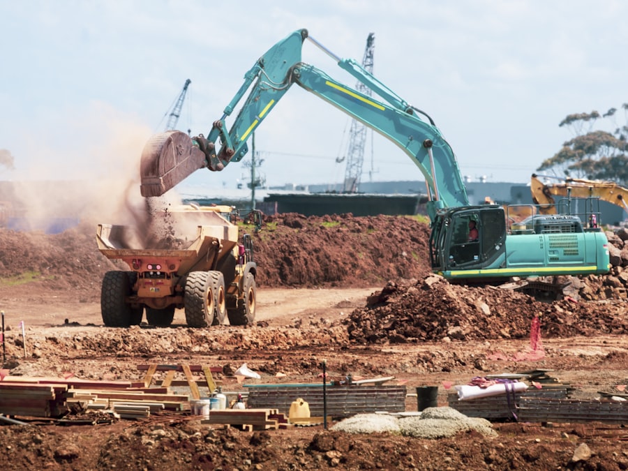 A teal excavator dumping soil into a yellow dump truck at a construction site